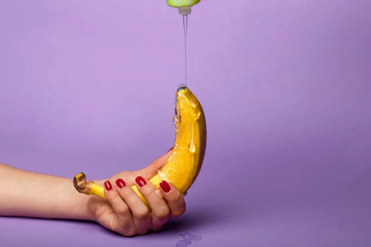 Hand with red nails holding banana while clear flavored lubricant or massage oil is poured over it on purple background